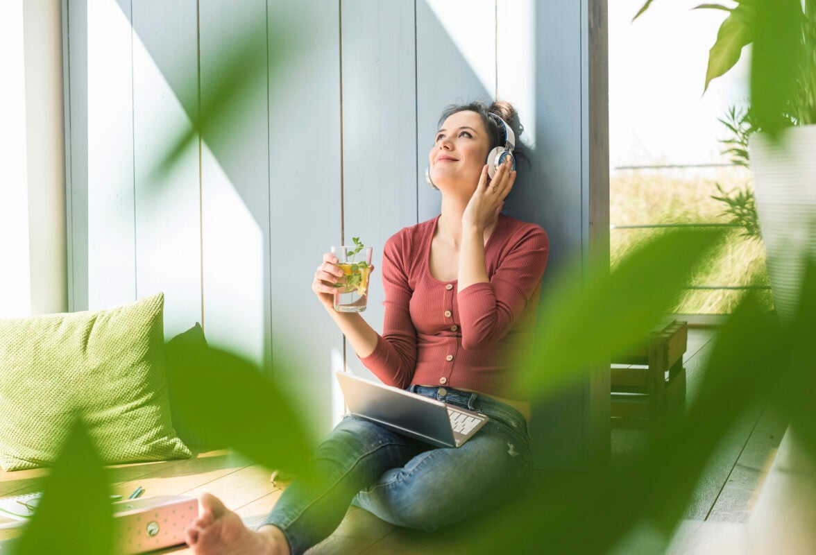 Smiling woman with headphones and laptop sitting at the window drinking clean water.